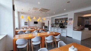 Un espace repas moderne avec des tables en bois et des chaises grises, une cuisine en arrière-plan et des suspensions jaunes.