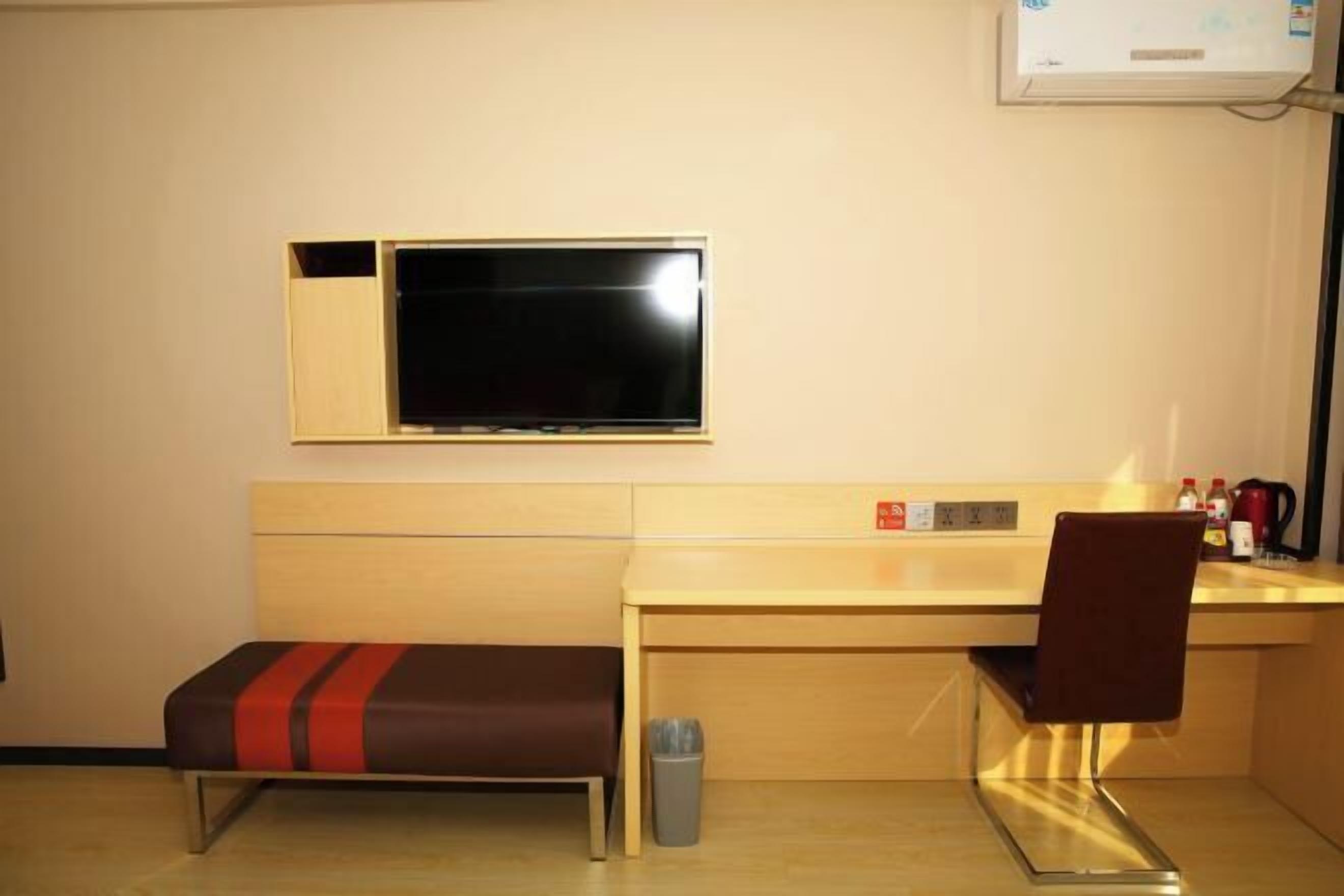 A hotel room with a bed, a desk, a chair, a TV, a trash bin, and a heater.