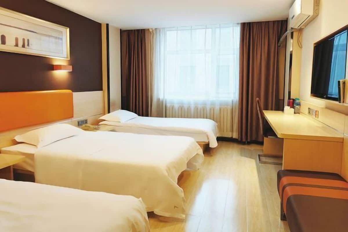 A hotel room with three beds, a desk, a chair, a window with curtains, and a wall-mounted air conditioner.