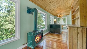 A room with a wood-burning stove, large windows, and a wooden ceiling.
