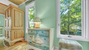 A room with a wooden cabinet, a dresser with a painted sailboat, a lamp, and a window with a view of trees.