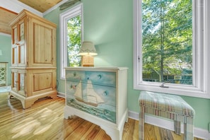 A room with a wooden cabinet, a dresser with a painted sailboat, a lamp, and a window with a view of trees.