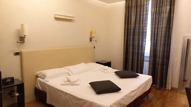 A hotel room with a bed, two bedside tables, a wall-mounted lamp, a window with curtains, and a radiator. - Stefano House Apartment Trevi Fountain  (Roma)
