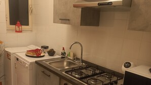 A compact kitchen with a gas stove, oven, and sink. There is a small refrigerator and a window with a red lantern. - Stefano House Apartment Trevi Fountain  (Roma)