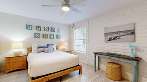 A bedroom with a bed, two bedside tables, a ceiling fan, a window with blinds, a wall-mounted picture, and a small table with a sign.