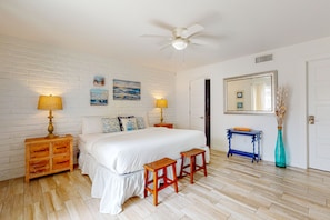 Quarto com parede de tijolos brancos, cama com roupa de cama branca, duas mesas de cabeceira com luminárias, uma mesinha lateral azul e um espelho na parede.