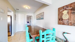 Un coin repas avec une table en bois et des chaises turquoise, un tableau représentant des voiliers et une pancarte « Bienvenue » accrochée au mur.