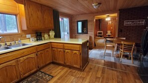 A wooden cabin interior with a kitchen featuring a double sink, wooden cabinets, and a dining area with a table and chairs. - Cottage #3 near a Creek & Pond on Mt. Lassen (Shingletown)