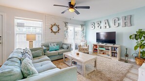 A living room with a sofa, coffee table, TV, bookshelf, and a ceiling fan. - Ground Floor Condo, Shared Pool, Grills and Boardwalk (Port Aransas)