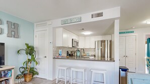 A modern kitchen with white cabinets, a stainless steel refrigerator, and a bar area with stools. - Ground Floor Condo, Shared Pool, Grills and Boardwalk (Port Aransas)
