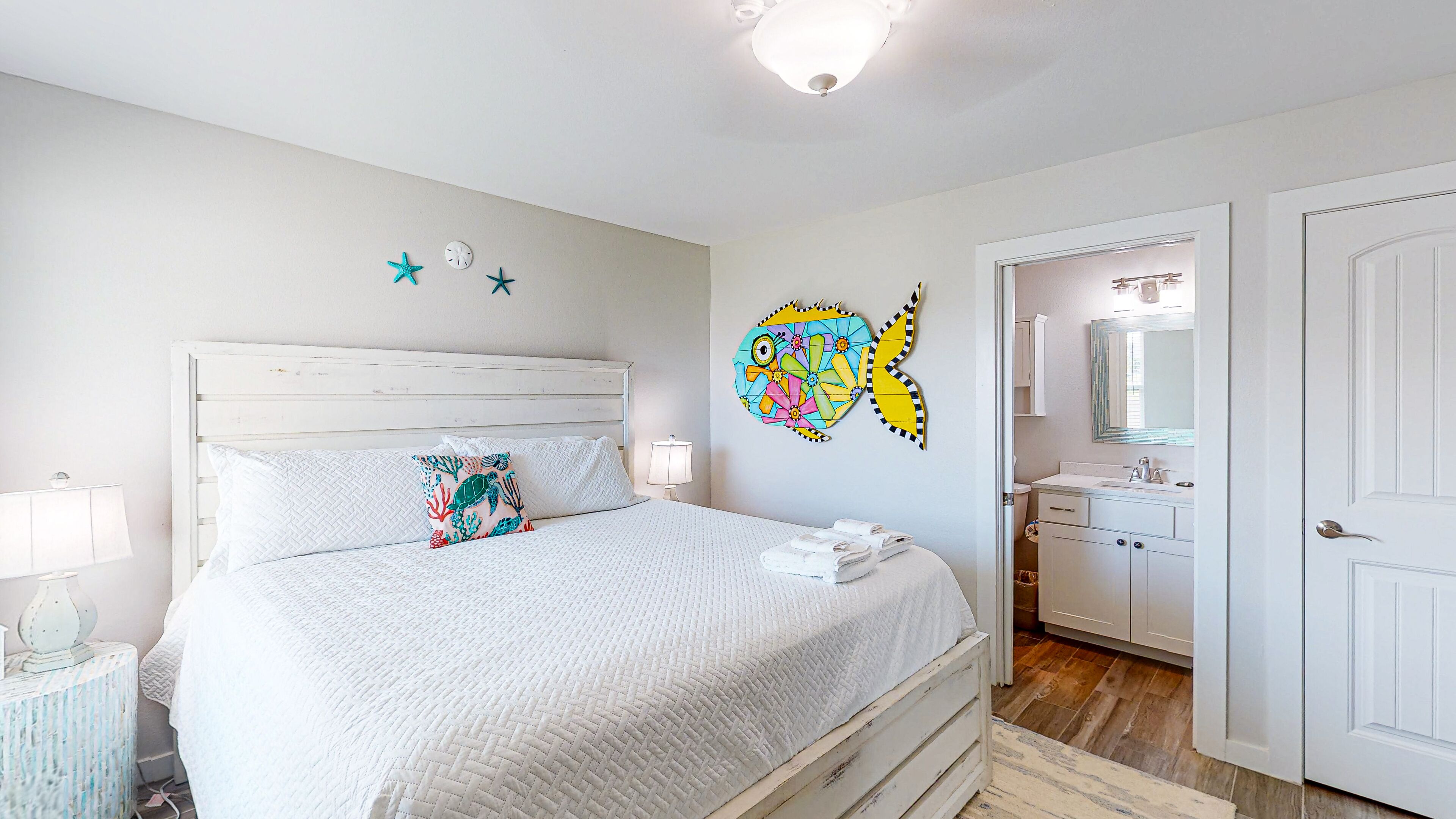 A bedroom with a bed, a nightstand, a lamp, a wall with decorative elements, and a bathroom visible through an open door.