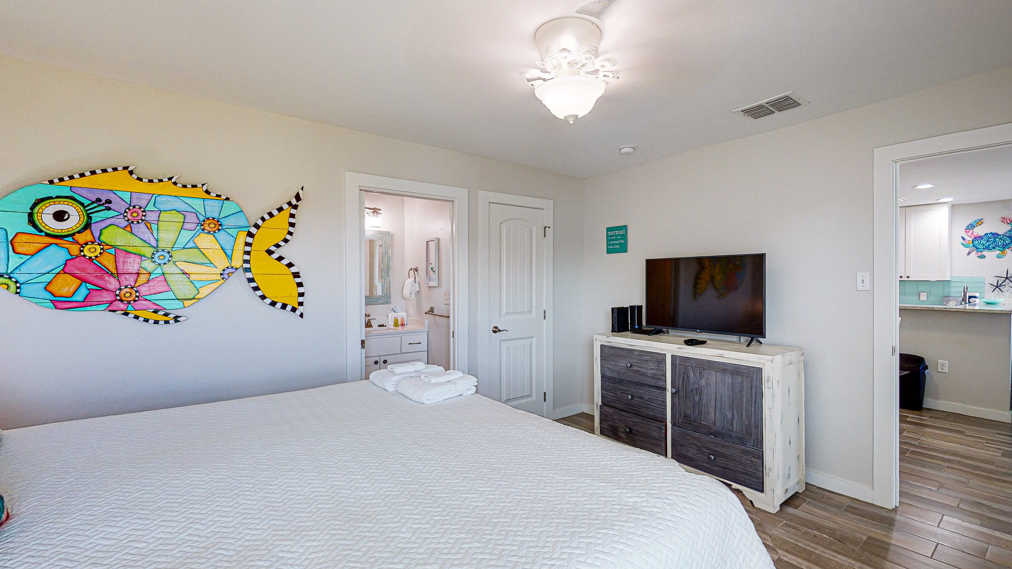 A bedroom with a bed, a TV on a dresser, a bathroom with a mirror, and a colorful wall decoration.