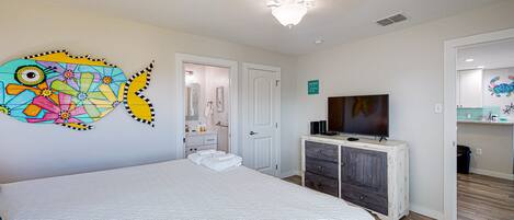 A bedroom with a bed, a TV on a dresser, a bathroom with a mirror, and a colorful wall decoration.
