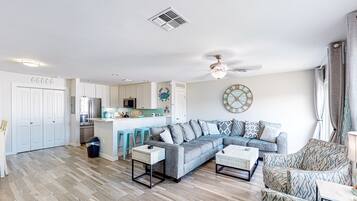 Una sala moderna con un sofá seccional, una mesa de centro y un televisor. Hay una zona de cocina con refrigerador, microondas y lavaplatos. La habitación tiene un ventilador de techo y un reloj de pared decorativo.