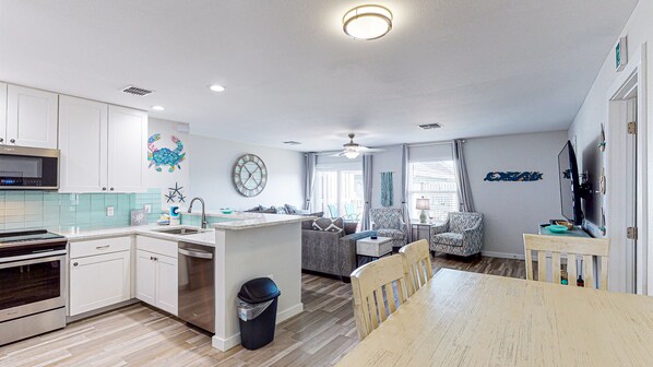 A modern kitchen with white cabinets, a microwave, and a dishwasher. There is a dining area with a wooden table and chairs. The living room has a sofa and a TV. - Second Floor Condo, Shared Pool, Grills and Boardwalk (Port Aransas)