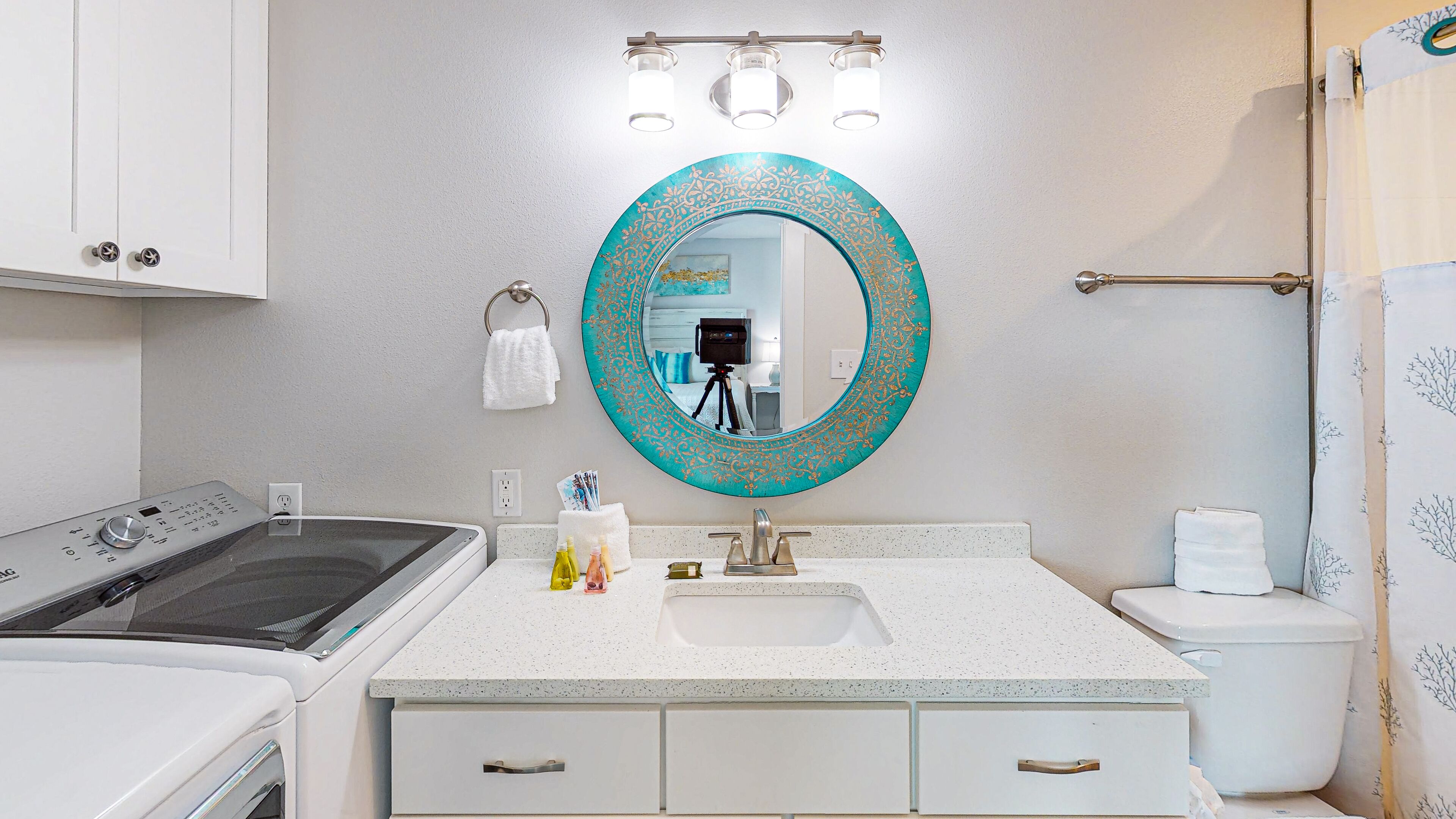 Un baño moderno con un lavamanos blanco, un espejo redondo con marco decorativo, un inodoro y una cortina de ducha con estampado de hojas.