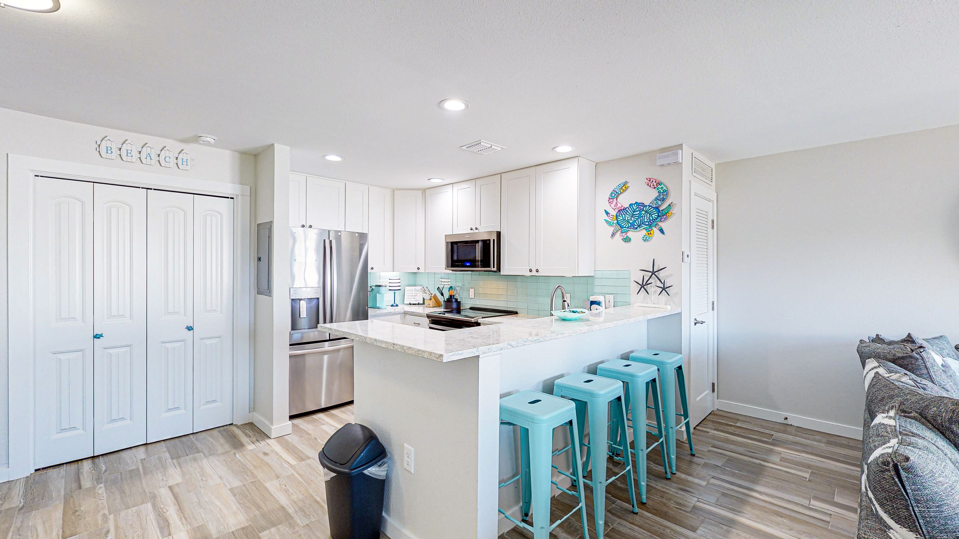 A modern kitchen with white cabinets, a stainless steel refrigerator, a microwave, and a sink. There are turquoise bar stools and a decorative turtle wall art piece.