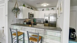 A kitchen with white cabinets, a stainless steel refrigerator, a microwave, and a coffee maker. There are two bar stools with woven seats. - Pet Friendly, Second Floor Condo, Shared Pool and Grill, Boardwalk (Port Aransas)