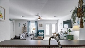 A modern kitchen with a central island, a dining area with a green chair, and a living room with a sofa and a television. - Pet Friendly, Second Floor Condo, Shared Pool and Grill, Boardwalk (Port Aransas)