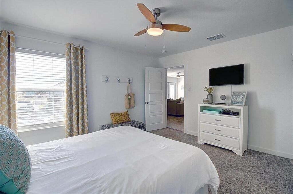 A bedroom with a bed, a ceiling fan, a TV, a dresser, and a door leading to another room.