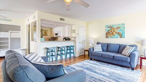 A living room with a sofa, a coffee table, and a kitchen area with a dining table and chairs. - Pet Friendly, Second Floor Condo, Shared Pool, Grills and Boardwalk (Port Aransas)