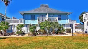 A two-story house with a blue exterior and white trim, featuring a front porch with a railing and a small garden with palm trees. - Pet Friendly, Second Floor Condo, Shared Pool, Grills and Boardwalk (Port Aransas)