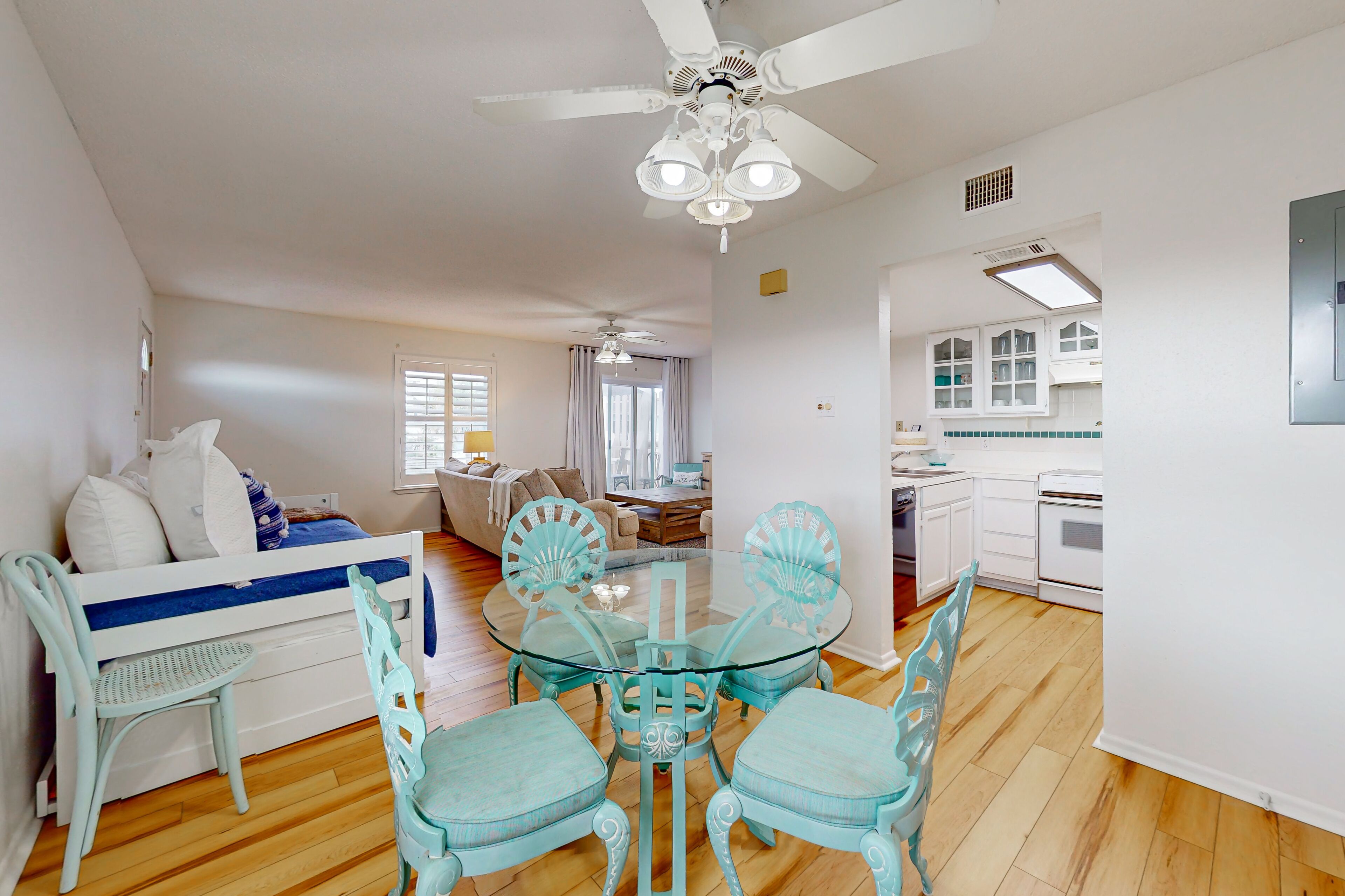 Un coin repas avec une table en verre et des chaises turquoise, un ventilateur de plafond et une cuisine en arrière-plan.