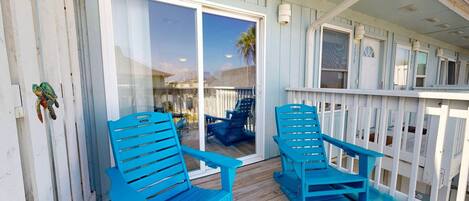 A covered porch with two blue Adirondack chairs, a glass door leading to a patio, and a white picket fence.