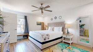 Ein Schlafzimmer mit Bett, Nachttischen, Deckenventilator, einem Fenster mit Vorhängen und Blick ins Freie.