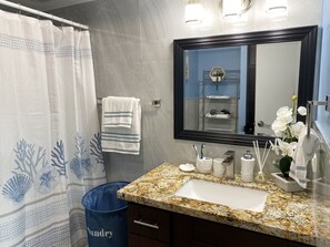 Een badkamer met een granieten wastafel, een spiegel, een handdoekhouder, een blauwe prullenbak en een douchegordijn met een blauw bloemenmotief.