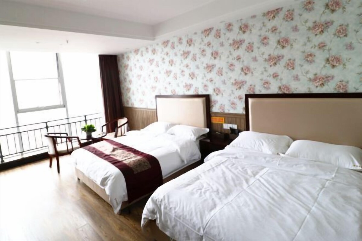 Habitación de hotel con dos camas, una mesita con una silla, una ventana con vistas y papel tapiz con motivos florales.