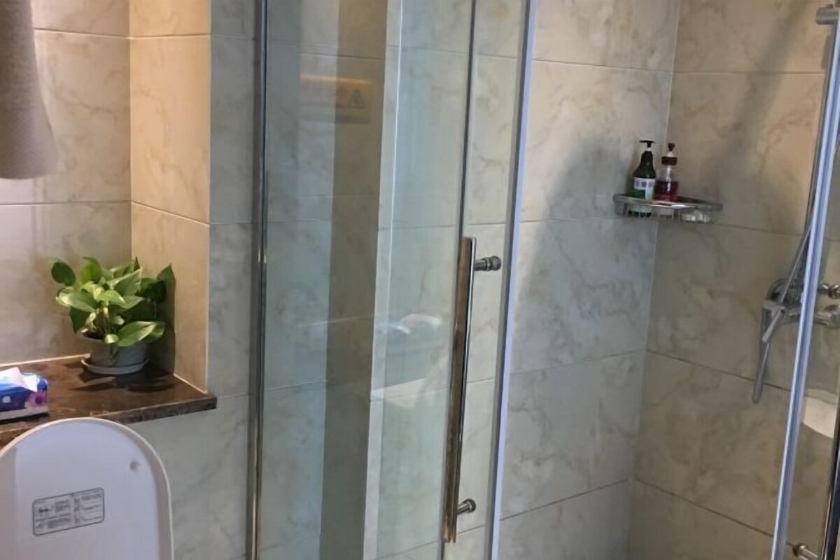 Un baño con cabina de ducha de vidrio, una planta en maceta y un dispensador de jabón en un estante.