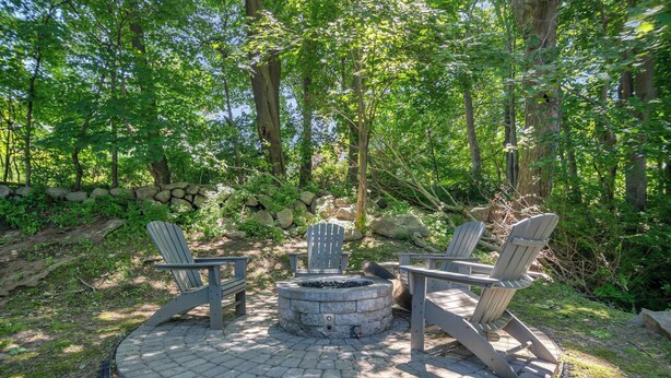 Un hogar circular con sillas Adirondack a su alrededor, ubicado en un patio empedrado rodeado por un muro de piedra y árboles.