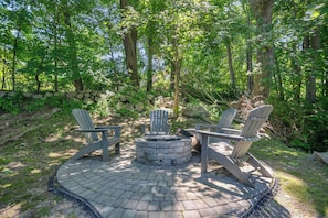 A circular fire pit with Adirondack chairs around it, set on a paved patio surrounded by a stone wall and trees.