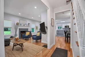 Un salon avec une cheminée, deux fauteuils bleus, une table basse et, au fond, une cuisine.
