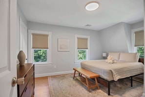 A bedroom with a bed, two windows, a bench, a dresser, and a lamp.