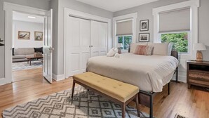 Quarto com cama, banqueta, mesa de cabeceira e janela com persianas.