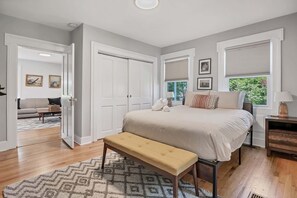 A bedroom with a bed, bench, bedside table, and a window with blinds. - Following Seas | Modern Charmer Walk to Downtown! (Mystic)
