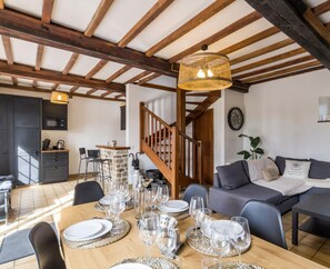 Un comedor con una mesa de madera dispuesta para una comida, una escalera y una sala de estar con un sofá y una planta.
