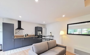 A modern kitchen with dark cabinets, a marble backsplash, and a built-in oven. There is a dining area with a table and chairs, and a grey sofa in the living area. - "La Boétie II" - 50 m2 de Charme Limoges Centre (Limoges)
