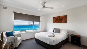Ein Schlafzimmer mit großem Fenster und Meerblick, einem Bett mit weißer Bettwäsche, einem Deckenventilator und einem kleinen Nachttisch.