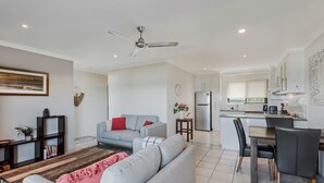 Ein modernes Wohnzimmer mit grauem Sofa, einem Holztisch und einem roten Zierkissen. Ein Essbereich mit einem schwarzen Stuhl und einem kleinen Tisch ist ebenfalls vorhanden. Im Hintergrund ist eine Küche mit weißen Schränken und einem Edelstahlkühlschrank zu sehen.