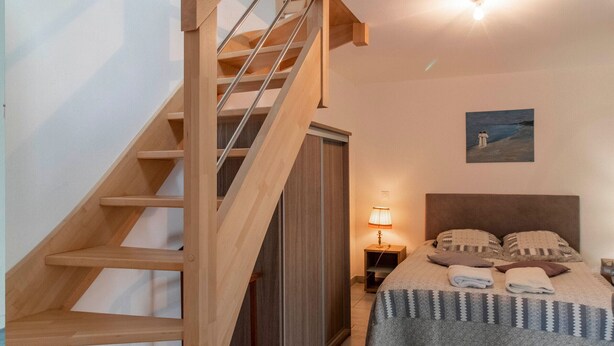 Un dormitorio con una escalera de madera, una cama con almohadas y una colcha, una mesita de noche con una lámpara y un cuadro en la pared.