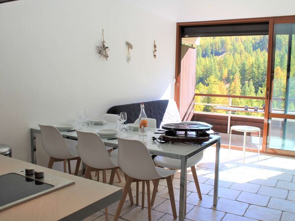 A dining area with a table set for two, a laptop, and a bottle of wine. There is a window with a view of a forest.