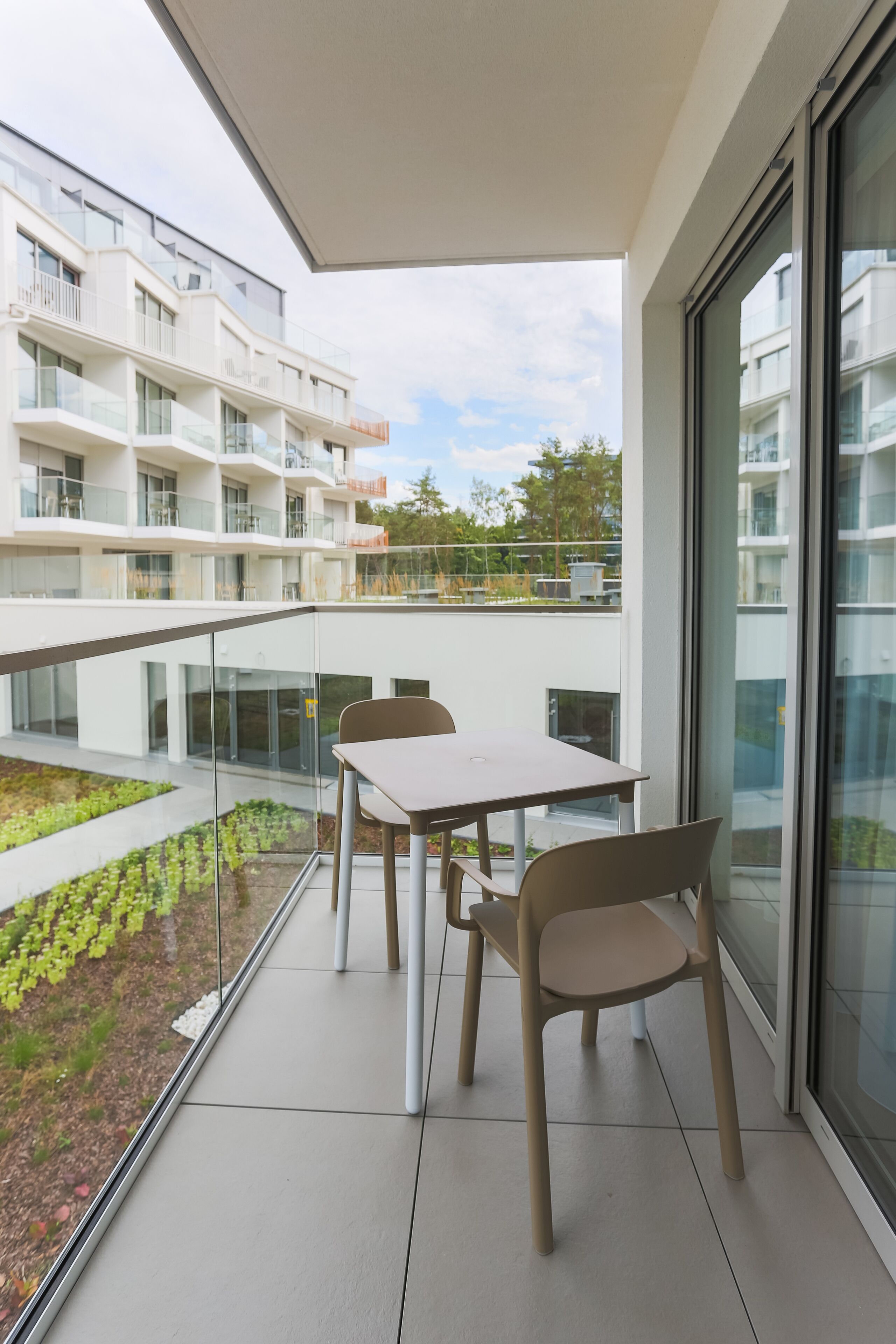 A balcony with a table and chairs, a glass railing, and a view of a modern building and greenery.
