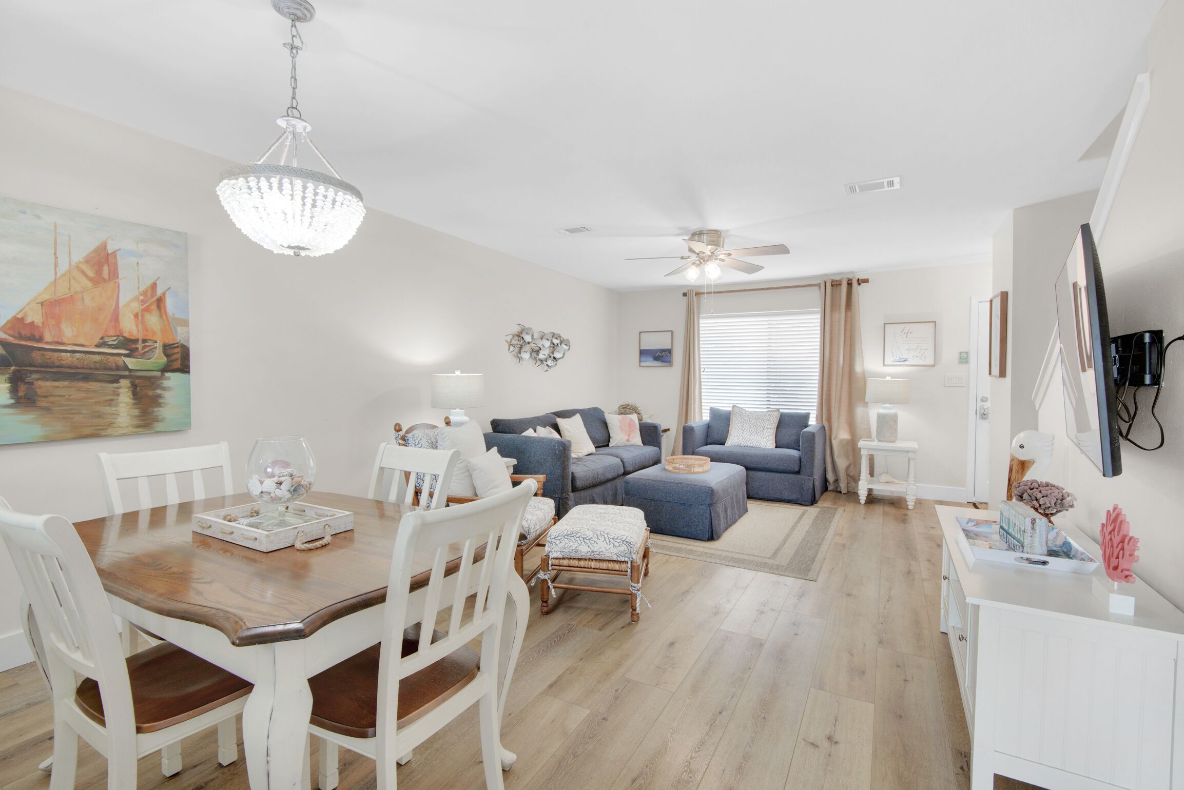 A spacious living room with a wooden dining table, white chairs, a blue sofa set, and a chandelier.