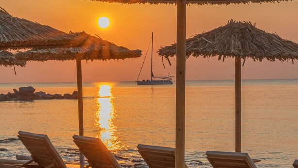 A beach with wooden lounge chairs and thatched umbrellas, a sailboat in the distance, and the sun setting over the water. - Cabana Blu Hotel & Suites (Kos)