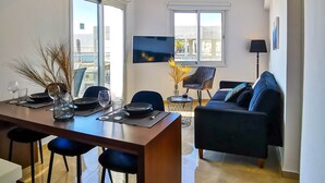 A modern living room with a sofa, a dining area with a table and chairs, and a TV mounted on the wall. - Phaedrus Living: Seaside Residence Lighthouse 70 (Paphos)