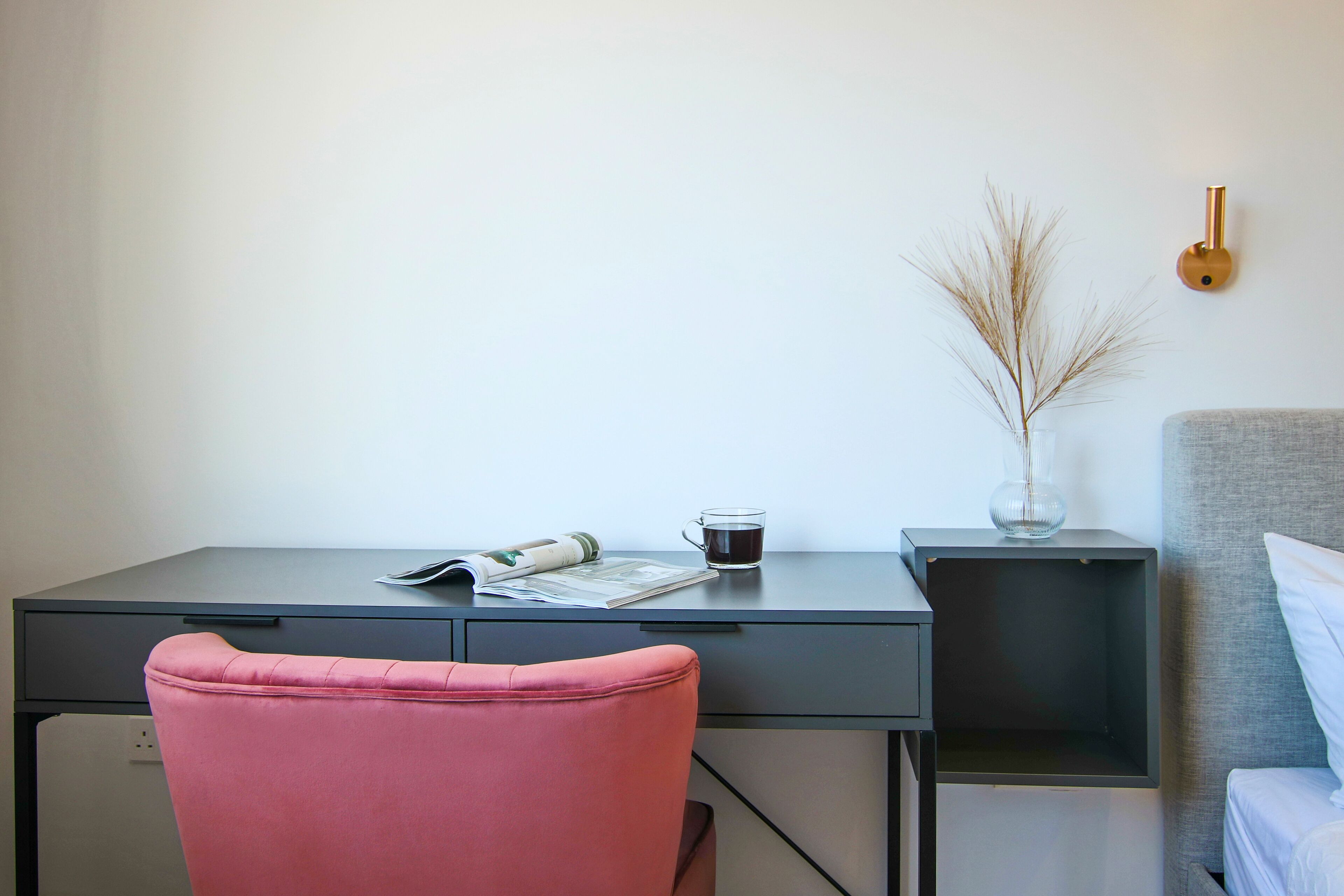 A modern room with a desk, a pink chair, a bedside table with a vase, and a bed with a grey headboard.