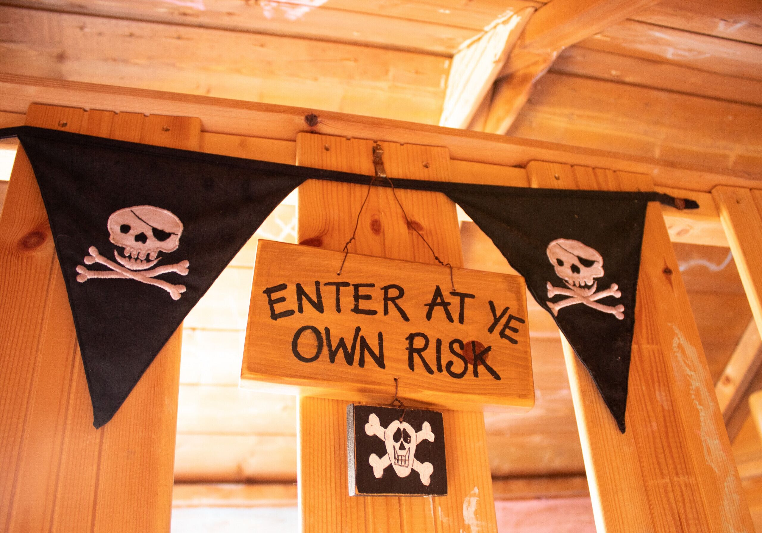 A wooden structure with a sign that reads 'ENTER AT YE OWN RISK' and two black triangular flags with skull and crossbones symbols.